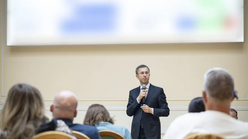 Промовець із мікрофоном перед екраном; частину слайда розмито у файлі джерела.